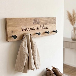 Könnte beinhalten: Eine rechteckige Wandgarderobe aus hellem Holz mit fünf schwarzen, runden Haken. Das Holz ist mit einem kleinen Regenbogensymbol und dem Schriftzug "Hanna & Elisa" in Schreibschrift graviert. Eine beige Jacke hängt an einem der Haken.
