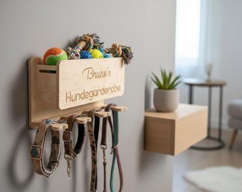 May include: A light-colored wooden wall-mounted organizer featuring a storage bin with the engraved text "Bruno's Hundegarderobe" and five hooks below. The bin holds colorful dog toys, while the hooks display various dog collars and leashes.