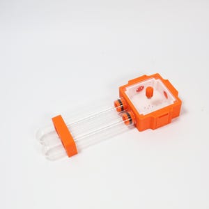 May include: An orange and clear ant farm. It features two clear tubes and a square orange habitat. The habitat has a clear top and a small orange knob. The tubes connect to the habitat with orange rings.
