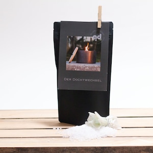 Könnte beinhalten: Ein schwarzer Beutel mit einem weißen Etikett, auf dem "Der Dochtwechsel" steht. Das Etikett zeigt ein Bild eines Kerzenhalters aus Keramik mit einer Flamme darin. Der Beutel liegt auf einer Holzoberfläche mit weißem Salz und einer getrockneten weißen Blume.