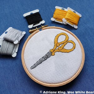 May include: A cross-stitch embroidery hoop with a yellow pair of scissors on a white background. The hoop is surrounded by spools of grey, black, and yellow embroidery floss.