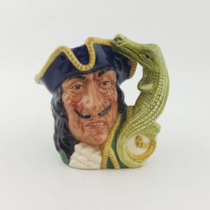 May include: A ceramic Captain Hook mug with a detailed face, a dark blue hat with gold trim, and a green crocodile handle. The mug has a cream-colored base and is painted with vibrant colours. A decorative item.