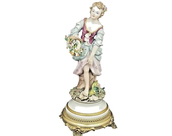 Puede incluir: Figura de porcelana de una mujer sosteniendo una cesta de flores. La mujer viste un vestido rosa y azul y se encuentra sobre una base decorativa con detalles dorados. La figura es un objeto decorativo.