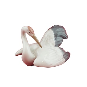 May include: A ceramic figurine of a crane in a resting pose. The bird has a white body with gray and white wings. The head and beak are a light brown color, with a touch of pink on the head.
