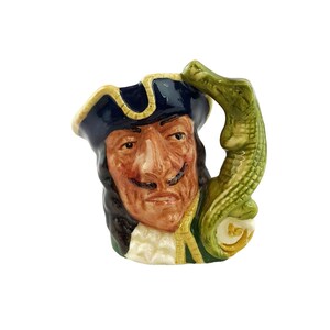 May include: A ceramic mug in the shape of a pirate's head, with a stern expression, a black mustache, and a tricorn hat. The handle is a green alligator. The mug has a cream-colored ruffled collar and gold trim. A collectible item.