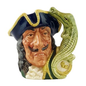 May include: A ceramic mug in the shape of a pirate's head, with a tricorn hat and a black mustache. A green alligator is wrapped around the side of the mug. The mug is painted in shades of brown, blue, and green, with a cream-colored collar.