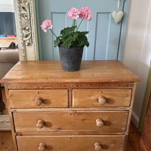 May include: A light brown wooden chest of drawers with two small top drawers and two full-width bottom drawers, each featuring round wooden knobs and keyholes. A dark grey pot containing a plant with pink flowers sits on the top surface.