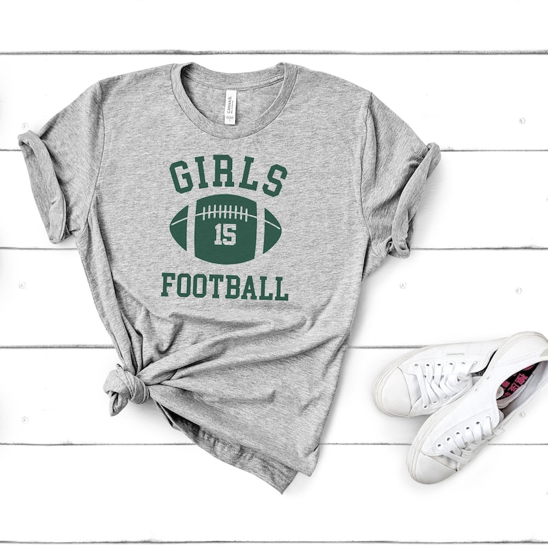 May include: Gray t-shirt with the text "GIRLS FOOTBALL" and a green football with the number 15 on it.
