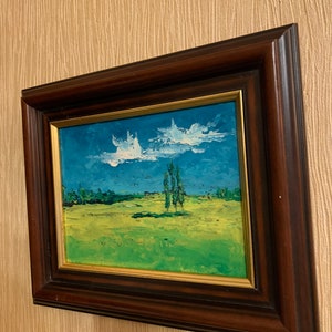 May include: A framed oil painting depicting a landscape scene with a field of green grass, a blue sky with white clouds, and three trees in the distance.