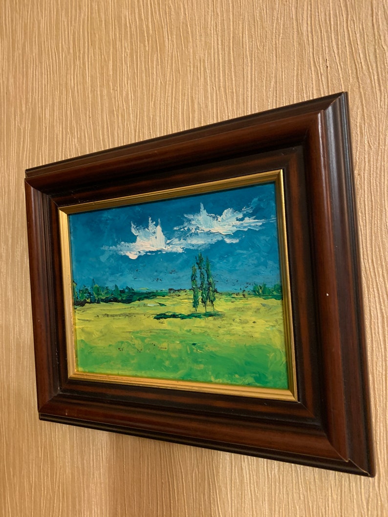 May include: A framed oil painting depicting a landscape scene with a field of green grass, a blue sky with white clouds, and three trees in the distance.