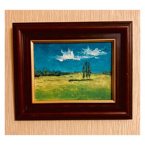 May include: A framed oil painting depicting a landscape with a field of green grass, a blue sky with white clouds, and three tall trees in the distance.