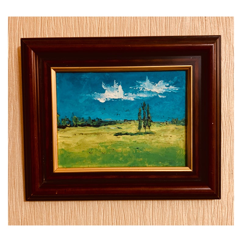 May include: A framed oil painting depicting a landscape with a field of green grass, a blue sky with white clouds, and three tall trees in the distance.