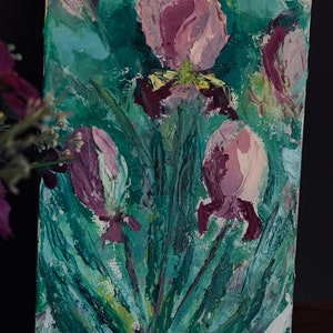 May include: A painting of purple irises with green leaves on a white canvas. The flowers are painted in a thick, textured style.