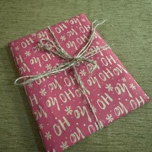 Puede incluir: Un regalo envuelto en papel rojo con las palabras "HO HO HO" y copos de nieve blancos. El paquete est&aacute; atado con hilo, creando un aspecto r&uacute;stico y festivo. Perfecto para regalos de Navidad o de vacaciones.