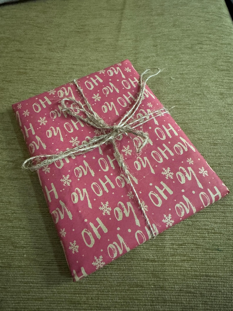Puede incluir: Un regalo envuelto en papel rojo con las palabras "HO HO HO" y copos de nieve blancos. El paquete est&aacute; atado con hilo, creando un aspecto r&uacute;stico y festivo. Perfecto para regalos de Navidad o de vacaciones.