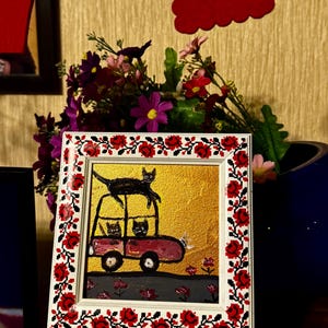 May include: A framed painting of a red car with three black cats inside. The car is on a gold background with red flowers. The frame has a white background with a red and black floral pattern.