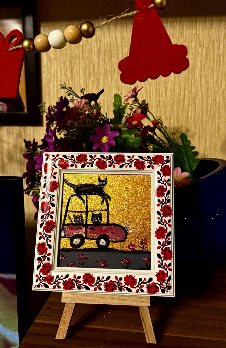 May include: A framed painting of a red car with three black cats inside. The car is on a gold background with red flowers. The frame has a white background with a red and black floral pattern.