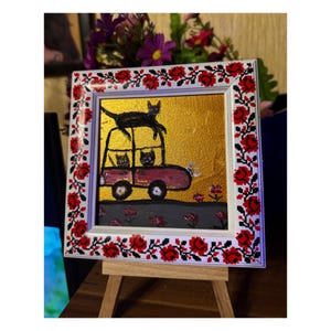 May include: A framed painting of a red car with three black cats inside and one black cat on top of the car. The car is on a gray road with pink flowers. The background is gold.