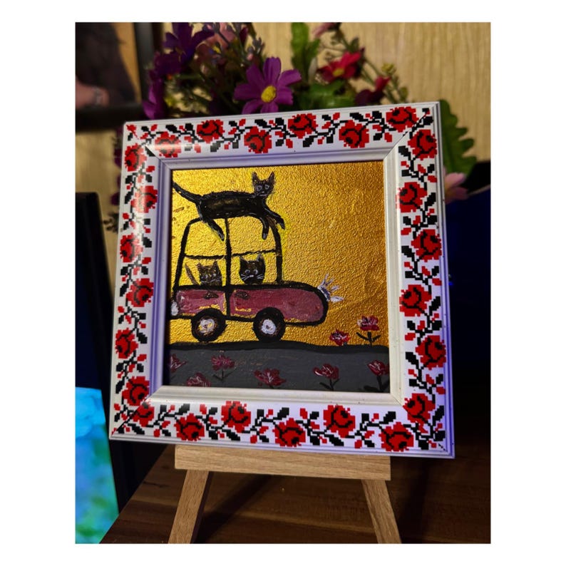 May include: A framed painting of a red car with three black cats inside and one black cat on top of the car. The car is on a gray road with pink flowers. The background is gold.