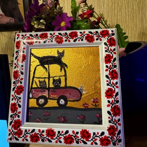 May include: A framed painting of a red car with three black cats inside. The car is on a gold background with pink flowers. The frame has a red and white floral design.