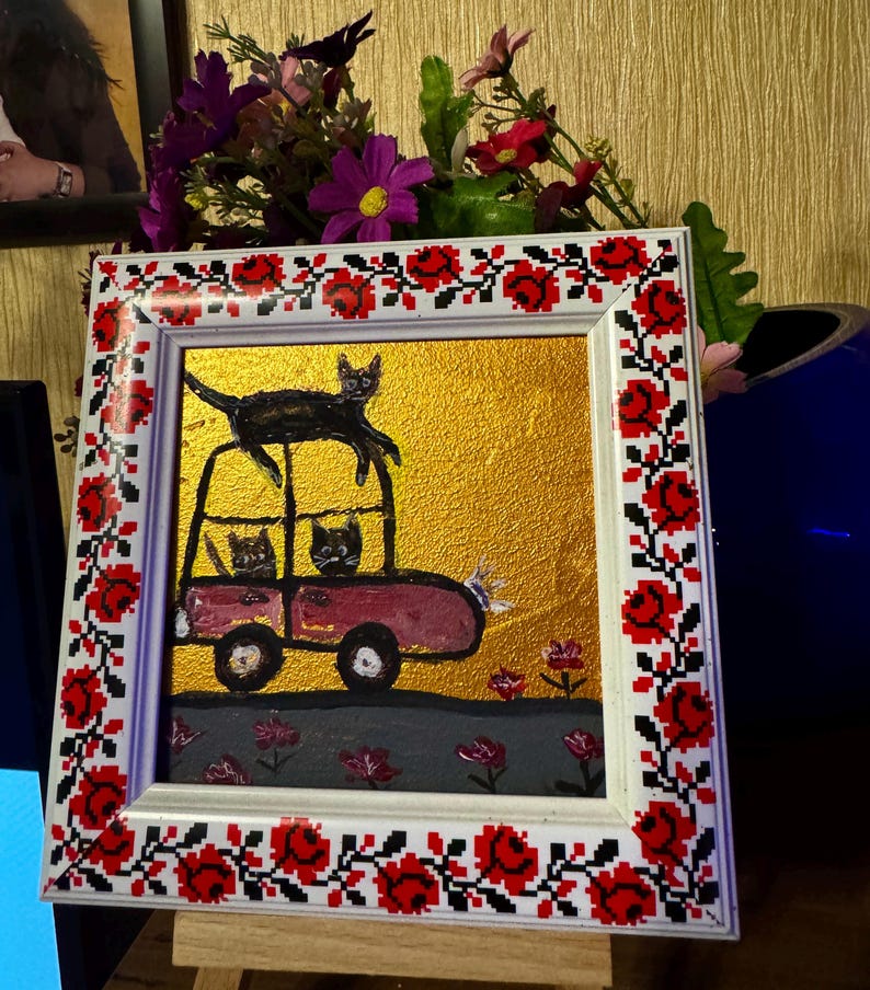 May include: A framed painting of a red car with three black cats inside. The car is on a gold background with pink flowers. The frame has a red and white floral design.