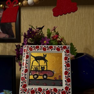 May include: A framed painting of three black cats in a pink car. The car is on a gold background with red flowers. The frame is decorated with a red and black floral pattern.