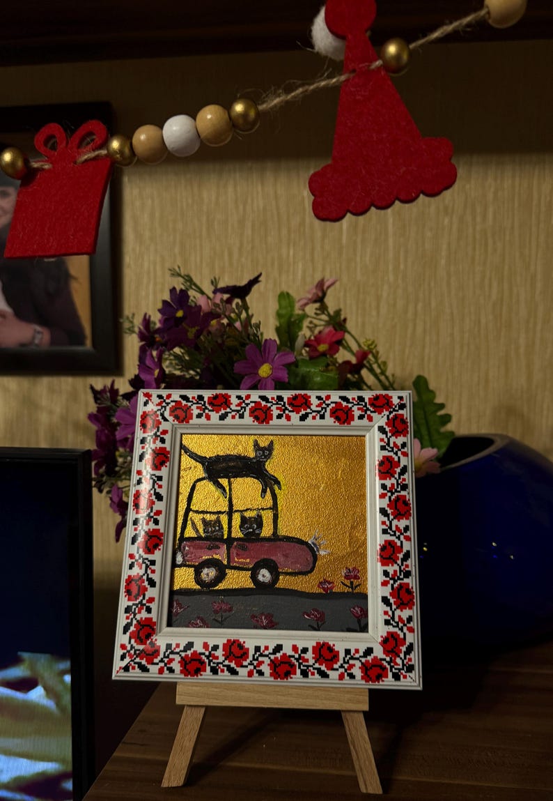 May include: A framed painting of three black cats in a pink car. The car is on a gold background with red flowers. The frame is decorated with a red and black floral pattern.