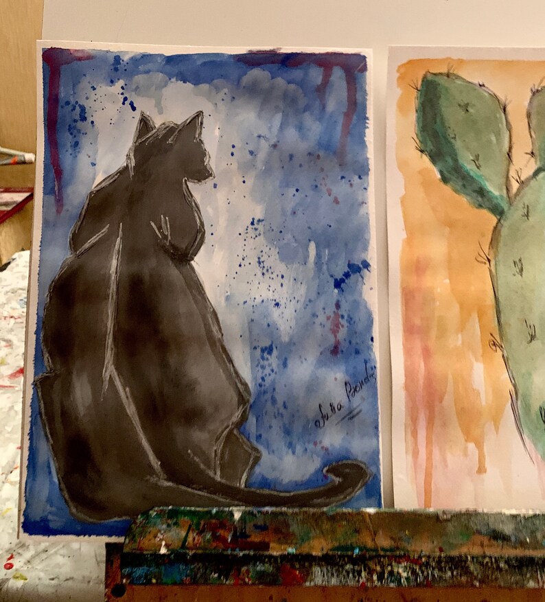 May include: A watercolor painting of a black cat sitting on a blue background with white splatters. The cat is facing away from the viewer. The artist's signature, "shura pachidi", is in the bottom right corner.