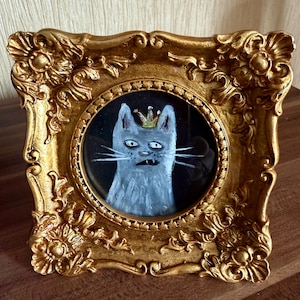 以下が含まれることがあります： 王冠をかぶった猫の額装絵画。猫はグレーと白の色合いで描かれ、背景は黒です。フレームは装飾的で金色です。