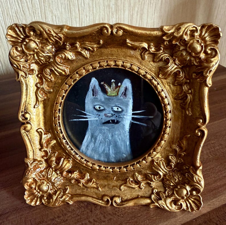 以下が含まれることがあります： 王冠をかぶった猫の額装絵画。猫はグレーと白の色合いで描かれ、背景は黒です。フレームは装飾的で金色です。