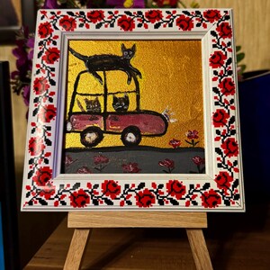 May include: A framed painting of a red car with three black cats inside and one black cat on top of the car. The car is driving on a gray road with red flowers. The frame has a white background with a red and white floral pattern.