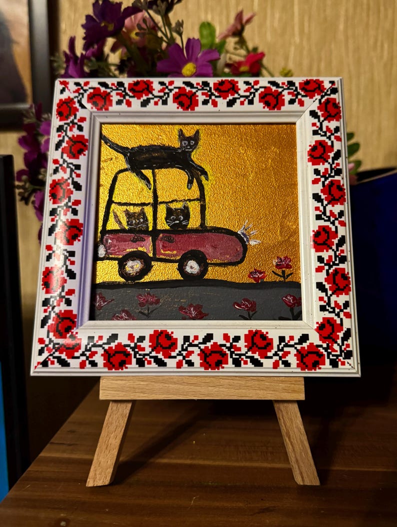 May include: A framed painting of a red car with three black cats inside and one black cat on top of the car. The car is driving on a gray road with red flowers. The frame has a white background with a red and white floral pattern.