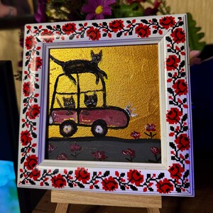May include: A framed painting of a pink car with three black cats on top of it. The car is driving on a gray road with pink flowers on either side. The background is gold. The frame is white with a red and black floral design.