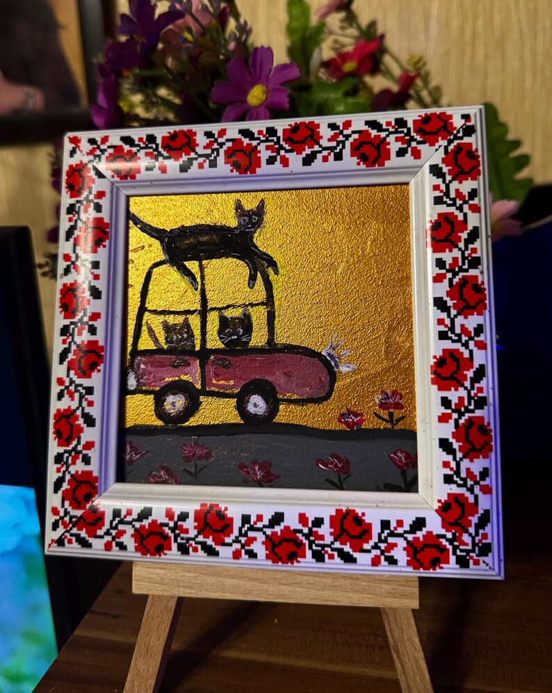 May include: A framed painting of a pink car with three black cats on top of it. The car is driving on a gray road with pink flowers on either side. The background is gold. The frame is white with a red and black floral design.