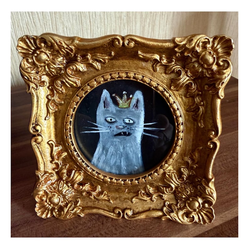 以下が含まれることがあります： 黒い背景に、金の王冠をかぶった灰色の猫の額装された絵。 猫は驚いた表情をしており、ひげと歯が見えます。 豪華な金のフレームがヴィンテージ感を加えています。