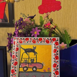 May include: A framed painting of three black cats riding in a pink car on a yellow background. The frame has a red and white embroidered border.