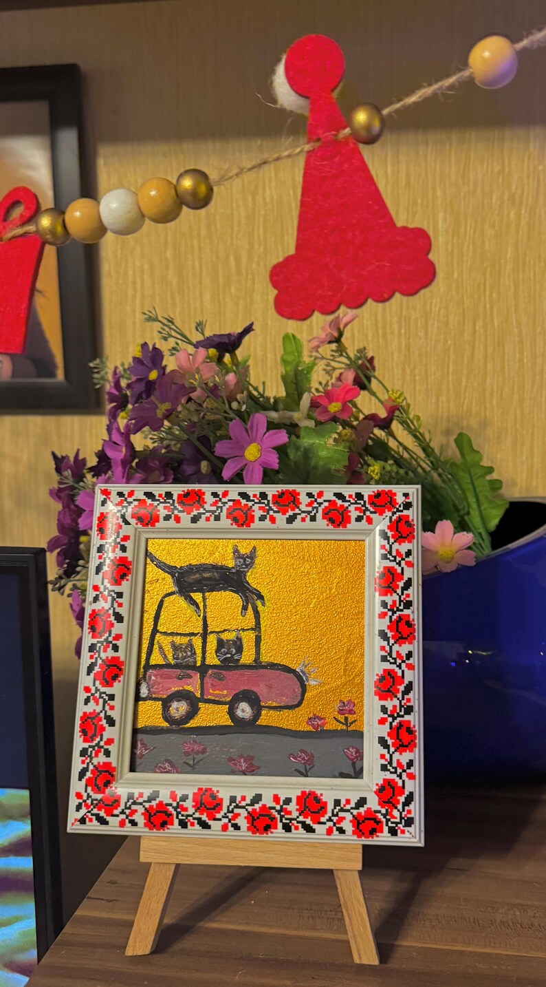 May include: A framed painting of three black cats riding in a pink car on a yellow background. The frame has a red and white embroidered border.
