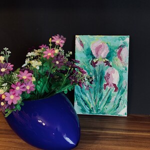 May include: A blue ceramic vase with a flower arrangement of purple and white flowers. A painting of pink flowers on a green background is on a wooden surface behind the vase.