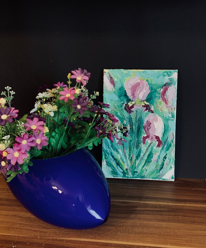May include: A blue ceramic vase with a flower arrangement of purple and white flowers. A painting of pink flowers on a green background is on a wooden surface behind the vase.
