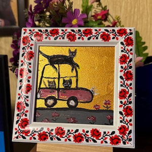 May include: A framed painting of a pink car with three black cats inside and one black cat on top of the car. The car is driving on a gray road with green grass and red flowers. The frame has a white background with a red and black floral pattern.