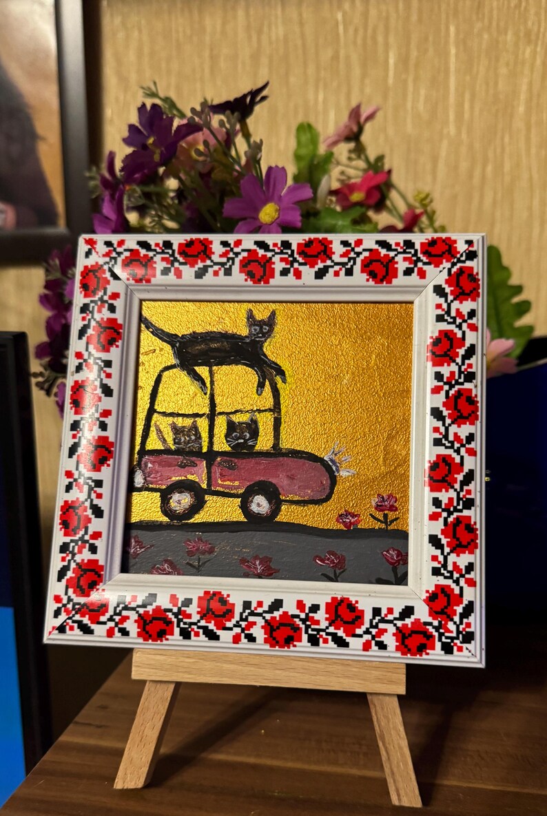 May include: A framed painting of a pink car with three black cats inside and one black cat on top of the car. The car is driving on a gray road with green grass and red flowers. The frame has a white background with a red and black floral pattern.