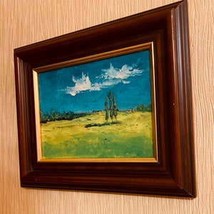 May include: A framed painting of a landscape with a blue sky, white clouds, green grass, and a few trees. The frame is dark brown wood with a gold trim.
