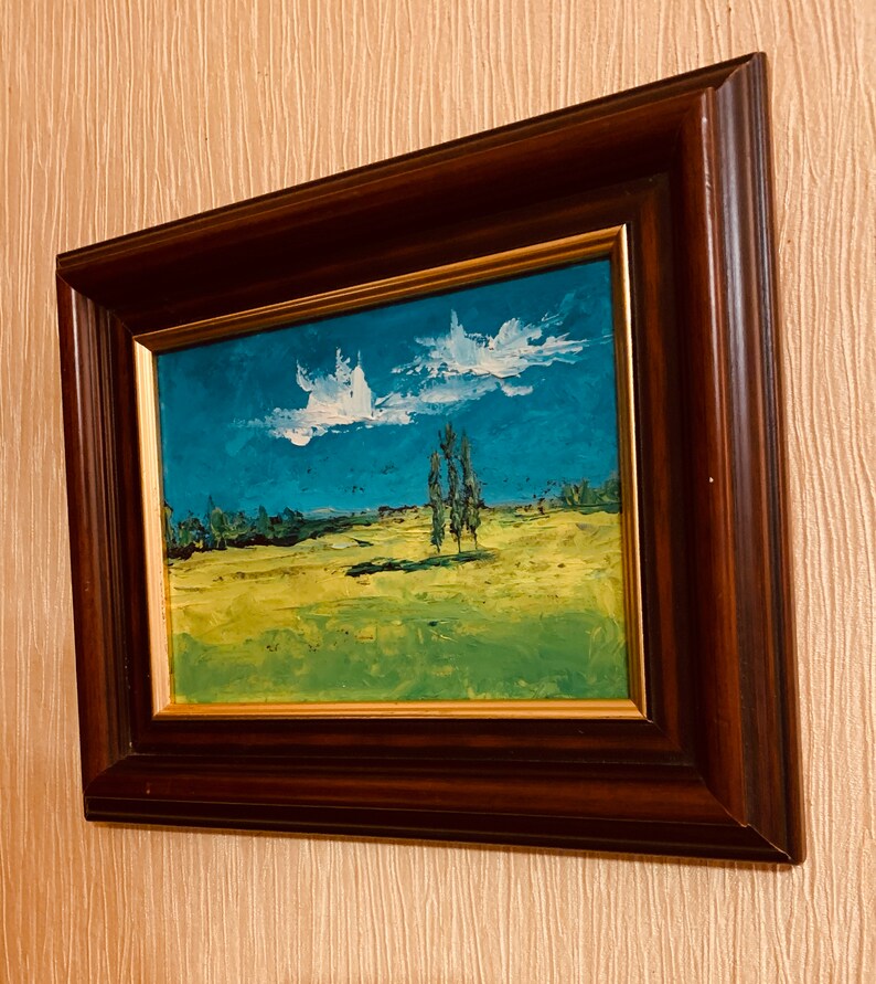 May include: A framed painting of a landscape with a blue sky, white clouds, green grass, and a few trees. The frame is dark brown wood with a gold trim.