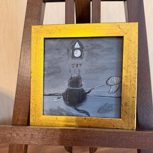 Puede incluir: Una pintura caprichosa en un marco dorado representa un gato negro con una red de mariposas, alcanzando un reloj. El fondo es una escena en escala de grises con una torre del reloj. La obra de arte se exhibe en un caballete de madera.