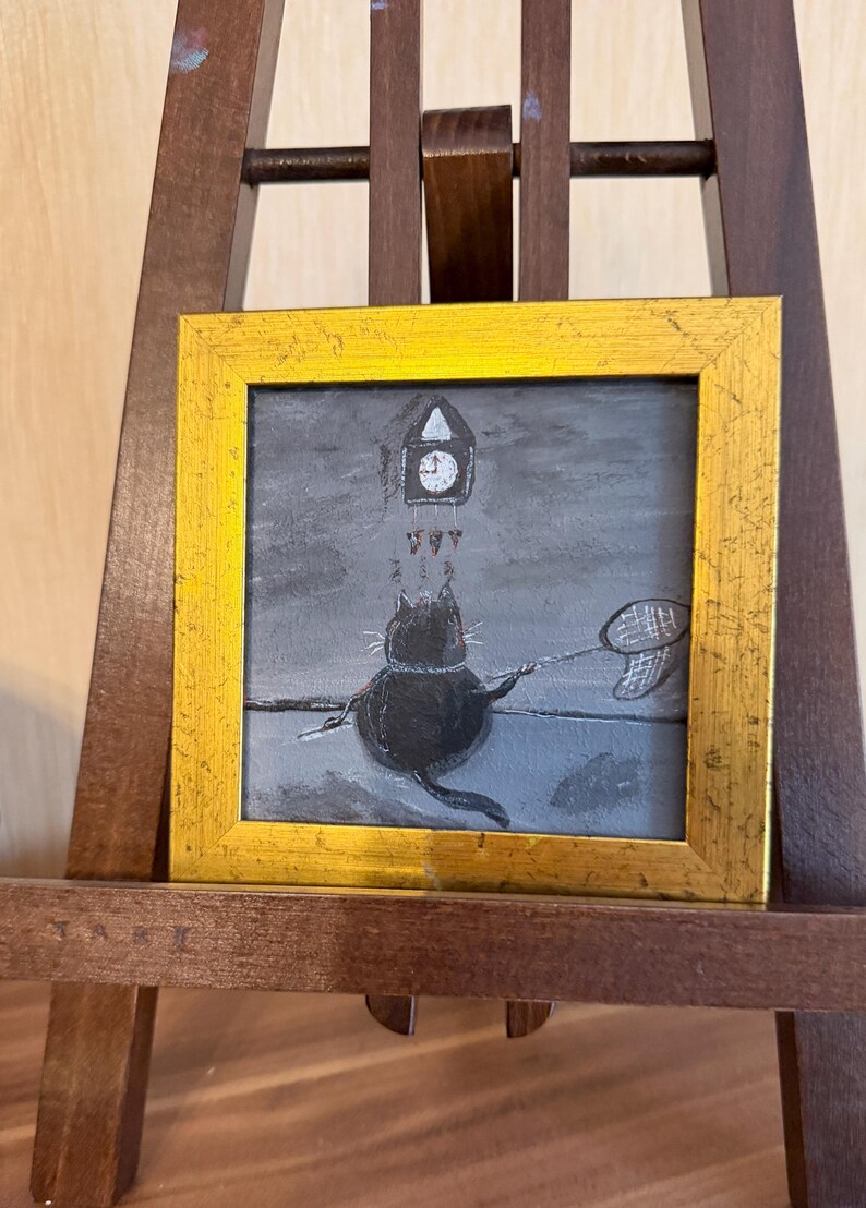 Puede incluir: Una pintura caprichosa en un marco dorado representa un gato negro con una red de mariposas, alcanzando un reloj. El fondo es una escena en escala de grises con una torre del reloj. La obra de arte se exhibe en un caballete de madera.