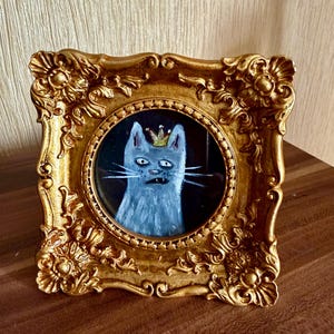 以下が含まれることがあります： 王冠をかぶった猫の額入り絵画。猫はグレーと白で描かれ、金の王冠が付いています。フレームは装飾的で金色、正方形です。絵は黒い背景に描かれています。