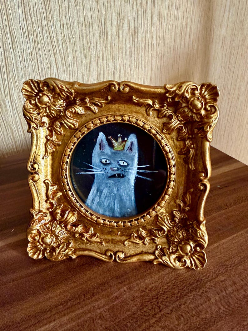 以下が含まれることがあります： 王冠をかぶった猫の額入り絵画。猫はグレーと白で描かれ、金の王冠が付いています。フレームは装飾的で金色、正方形です。絵は黒い背景に描かれています。