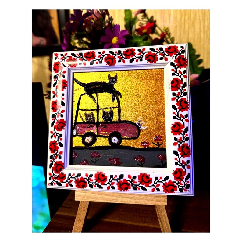 May include: A framed painting of a red car with three black cats inside and one black cat on top of the car. The car is on a gray road with red flowers. The frame is white with a red and black floral design.