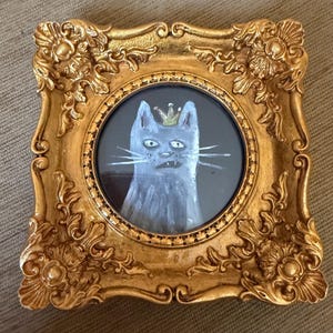 以下が含まれることがあります： 王冠をかぶった猫の額装絵画。猫はグレーと白の色合いで描かれ、金の王冠が描かれています。フレームは装飾的で金色、長方形です。