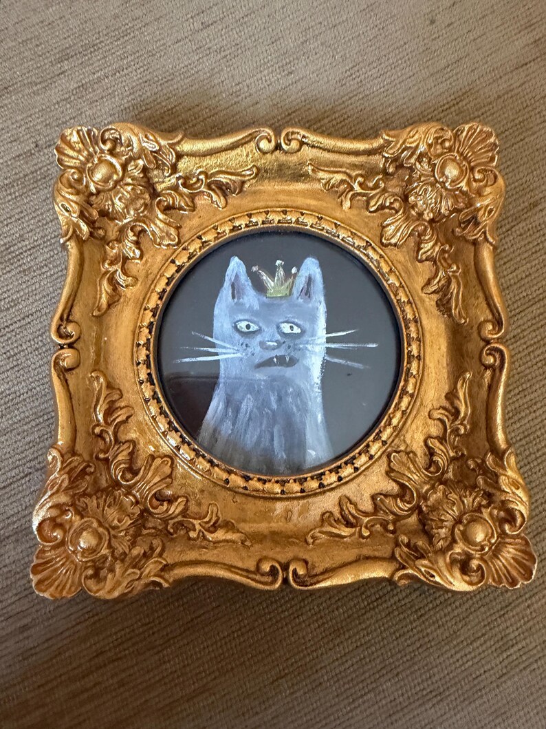 以下が含まれることがあります： 王冠をかぶった猫の額装絵画。猫はグレーと白の色合いで描かれ、金の王冠が描かれています。フレームは装飾的で金色、長方形です。